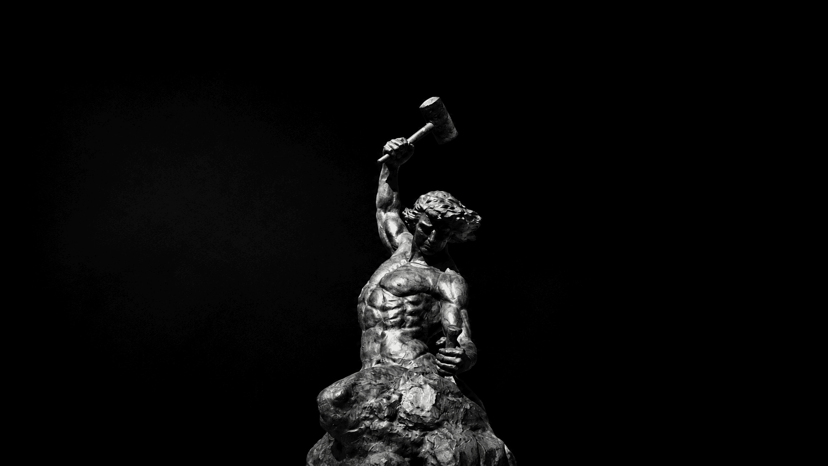 A sculpted figure raising a hammer and holding a chisel above stone.